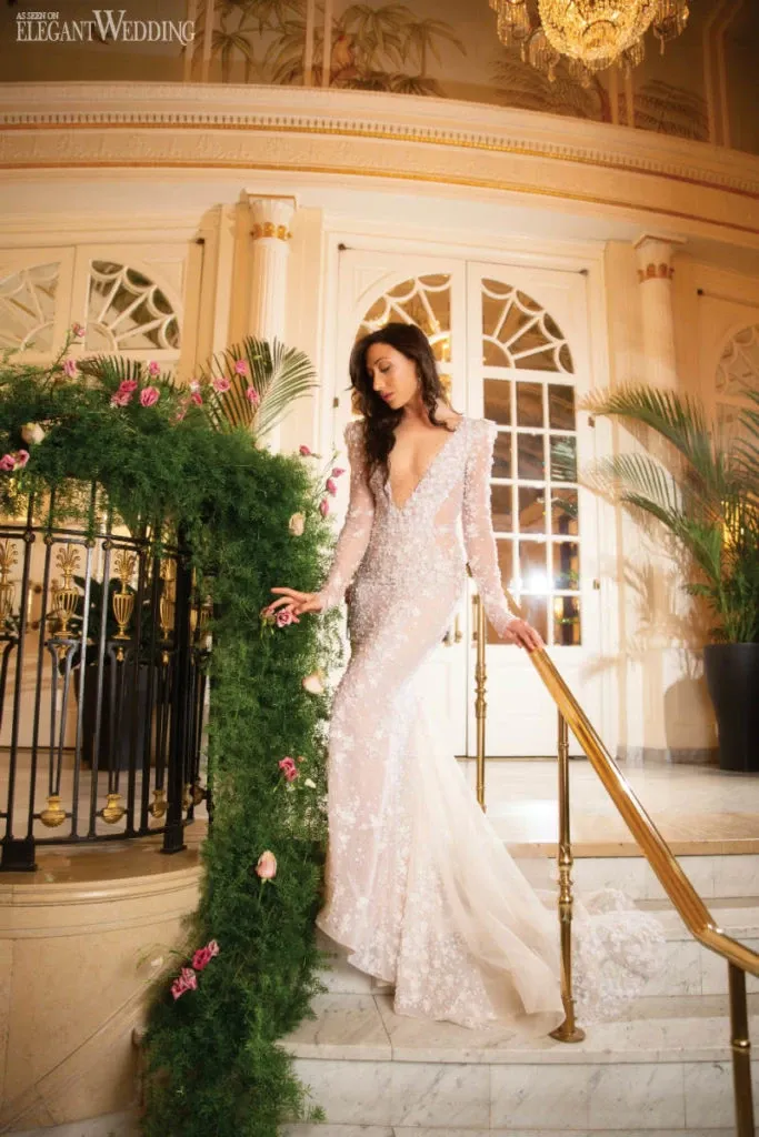 A bride descends an elegant marble staircase in a luxurious setting, showcasing a stunning wedding dress by White Montreal. The dress features intricate lace details and a gracefully flowing train that cascades down the steps. The long sleeves and deep neckline add a touch of sophistication. In the background, grand doors and opulent chandeliers enhance the luxurious atmosphere, while lush greenery adorned with delicate pink roses decorates the railing, adding a romantic touch to the wedding venue. This image highlights the exquisite design of White Montreal's wedding attire, perfectly fitting the elegant and refined theme of Mariage Elegant, Canada.