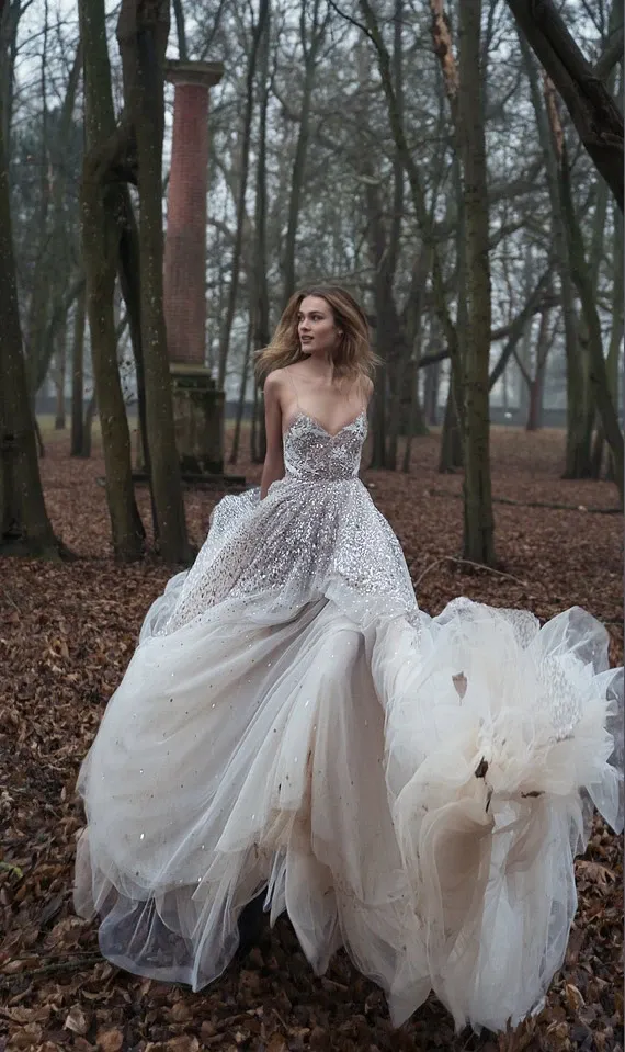 In a serene woodland setting, a bride wearing a stunning wedding dress by White Montreal, from the wedding dress category, stands elegantly amidst tall trees. The dress features a shimmering, intricately beaded bodice with a sweetheart neckline, seamlessly flowing into a voluminous tulle skirt that captures soft, natural light, enhancing its ethereal beauty. The bride's hair cascades down naturally, complementing the elegance of the gown, which is perfect for a sophisticated, fairy-tale wedding.