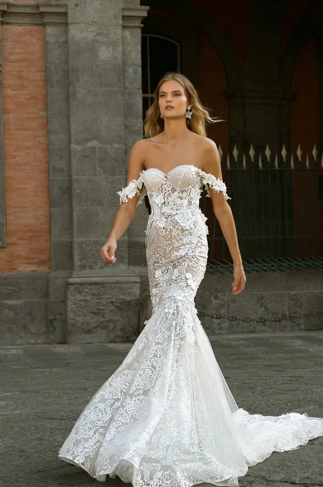 A bride models an elegant, off-the-shoulder lace wedding dress designed by White Montreal. The gown features a form-fitting silhouette with intricate lace detailing and delicate floral appliqués cascading down the bodice and sleeves. The skirt flows gracefully into a subtle train. This sophisticated wedding dress is perfectly suited for modern brides. The background shows an architectural setting, adding a classic touch to the scene, enhancing the elegance of the wedding dress category.