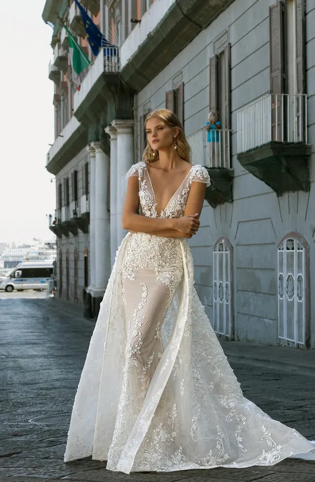 A bride elegantly poses in a stunning wedding dress by White Montreal, featured under the Wedding Dress category on Mariage Elegant, Canada. The gown is intricately designed with lace detailing, a deep V-neckline, and cap sleeves that accentuate its sophisticated style. A sheer overlay creates a flowing train, adding a touch of grace as it trails behind. The bride stands confidently on a cobblestone street, with a backdrop of historic European-style architecture. This image captures the timeless elegance that White Montreal brings to bridal fashion.