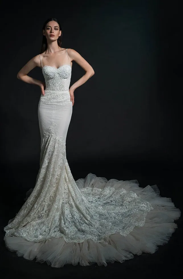 A bride is elegantly showcasing a wedding dress in this image from White Montreal, a vendor listed in the wedding dress category at Mariage Elegant, Canada. The dress features delicate lace detailing and a fitted bodice with thin straps. The gown flows into a dramatic, textured train that spreads gracefully on the floor. The background is a dark, neutral color, drawing attention to the intricate design and craftsmanship of the wedding dress.