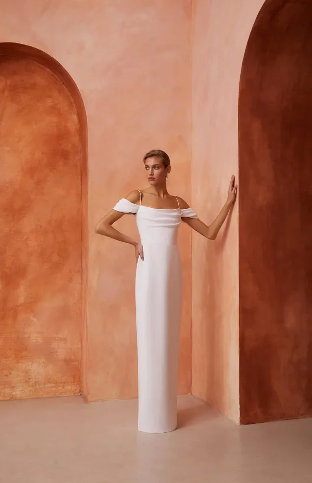 A bride stands elegantly against a warm-toned, textured arched background, wearing a sleek, minimalist white wedding dress designed by White Montreal. The gown, featured in the wedding dress category, showcases an off-the-shoulder design with thin straps and a floor-length, fitted silhouette, perfectly embodying a modern yet timeless bridal look. The setting, with its soft, earthy tones, enhances the sophistication and elegance of the dress, ideal for a wedding theme emphasizing simple yet refined aesthetics.