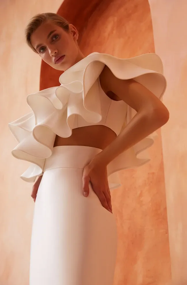 A bride stands elegantly in a sophisticated wedding dress by White Montreal, showcased in the Wedding Dress category. The dress features a striking, modern design with dramatic ruffled sleeves and a sleek, form-fitting silhouette. The fabric is pristine white, complementing the minimalistic yet bold fashion choice. The background features a warm terracotta arch, adding an artistic and stylish contrast to the scene, perfect for a contemporary wedding setting.