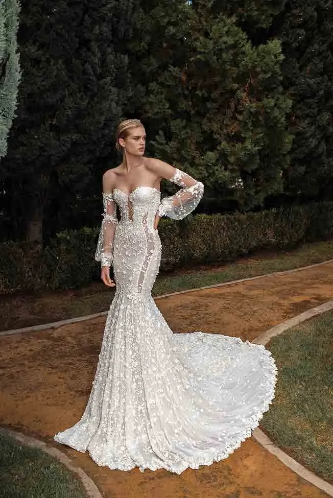 A bride is wearing an elegant wedding dress by White Montreal, featured in the wedding dress section of Mariage Elegant, Canada. The gown is an off-the-shoulder style with sheer, lace-embellished sleeves. The fitted bodice is adorned with intricate floral embroidery, extending to a beautiful, flowing train. The background features a serene garden setting, with lush greenery and a paved path, enhancing the romantic ambiance of the scene.