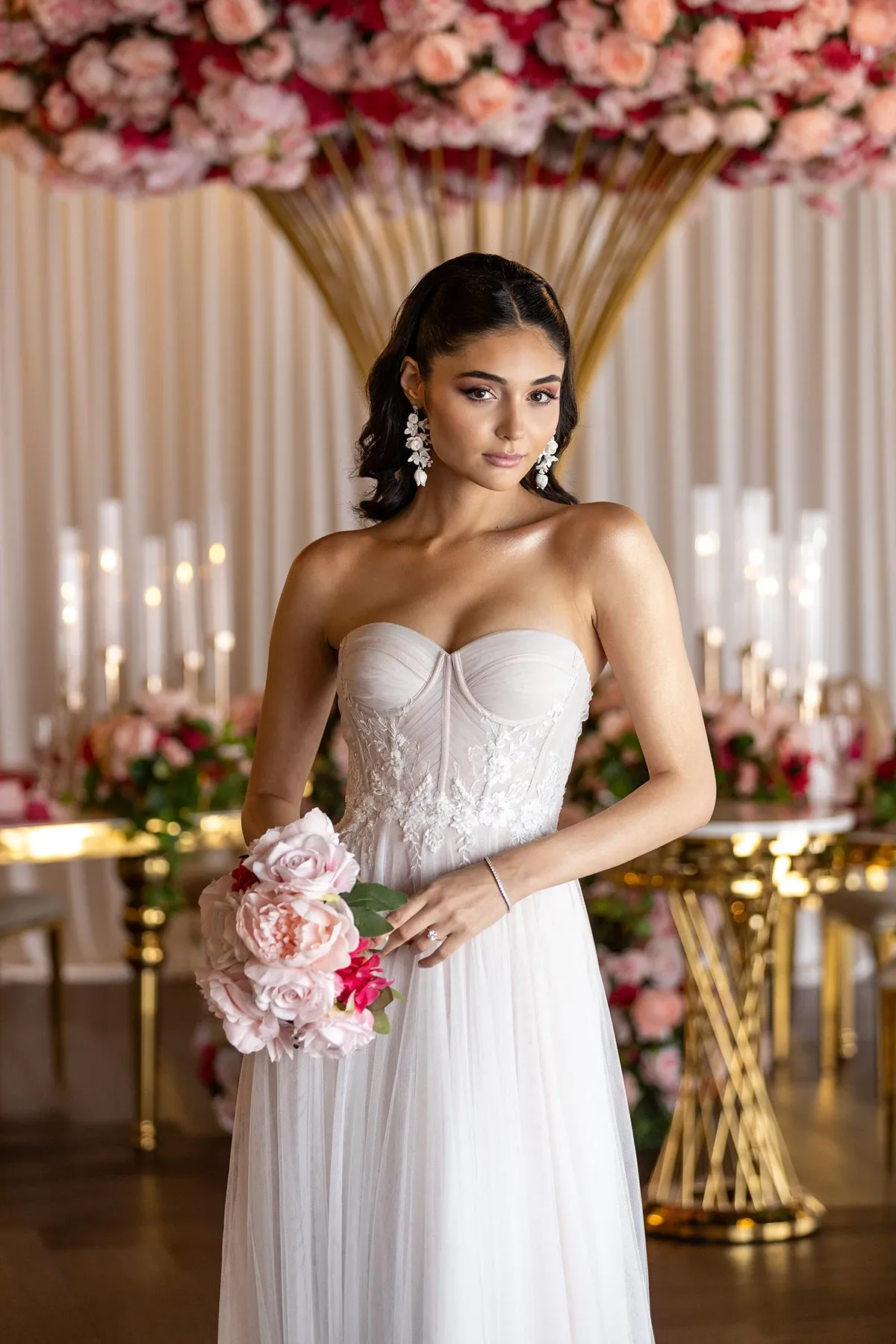 This image features a bride wearing an elegant wedding dress from Boutique MariClod, a vendor listed under the Wedding Dress category on Mariage Elegant in Canada. The strapless gown showcases a fitted bodice with intricate lace detailing, beautifully accentuating the bride's silhouette. She holds a delicate bouquet of soft pink and red roses, complementing the floral arrangements in the background. The setting includes an array of tall, white candles and a lush display of flowers, creating a romantic and luxurious atmosphere ideal for a wedding celebration.