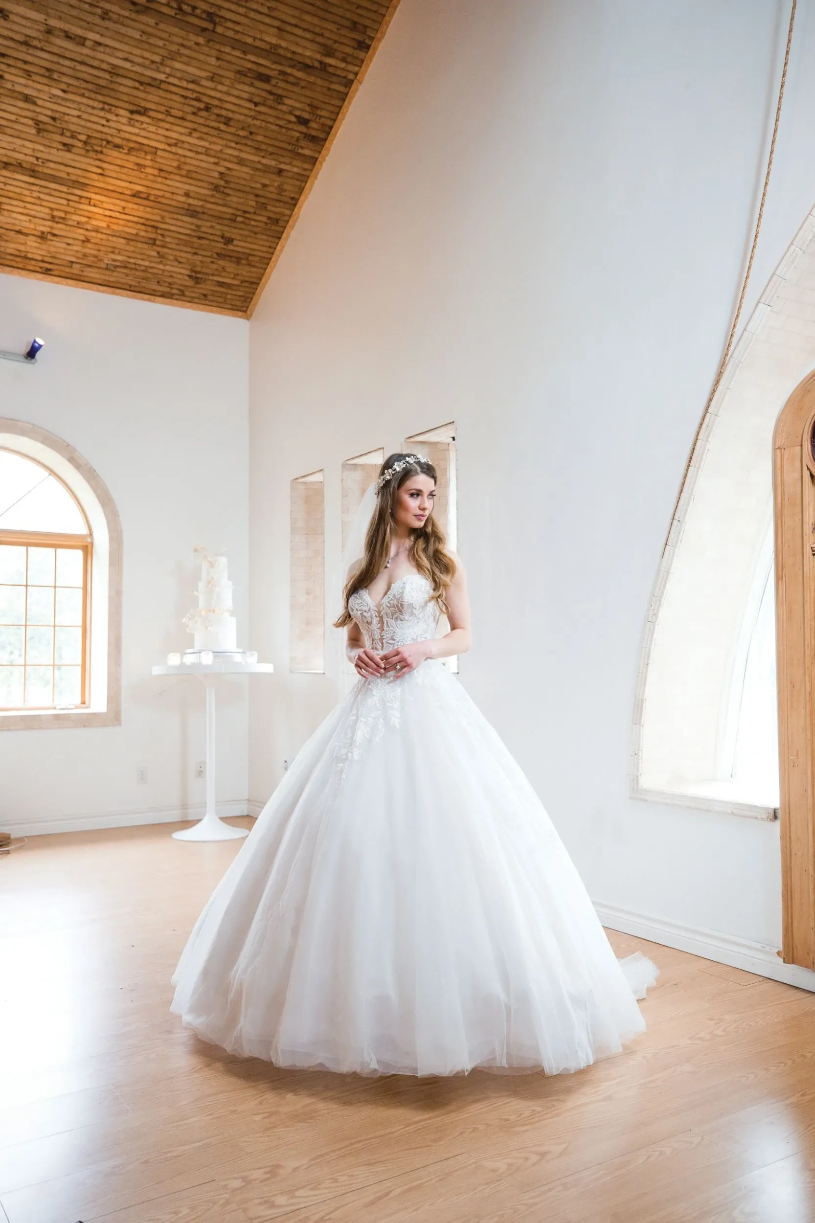 A bride stands gracefully in a beautifully lit room, wearing an elegant wedding gown from Boutique MariClod, categorized under Wedding Dress. The dress features an intricate lace bodice with delicate embroidery and a voluminous tulle skirt, creating a classic yet sophisticated bridal look. Natural light pours in through large, arched windows, illuminating the soft details of the gown. The room's wooden ceiling and minimalist decor enhance the romantic and serene ambiance, making it an ideal setting for showcasing this stunning dress by Boutique MariClod.