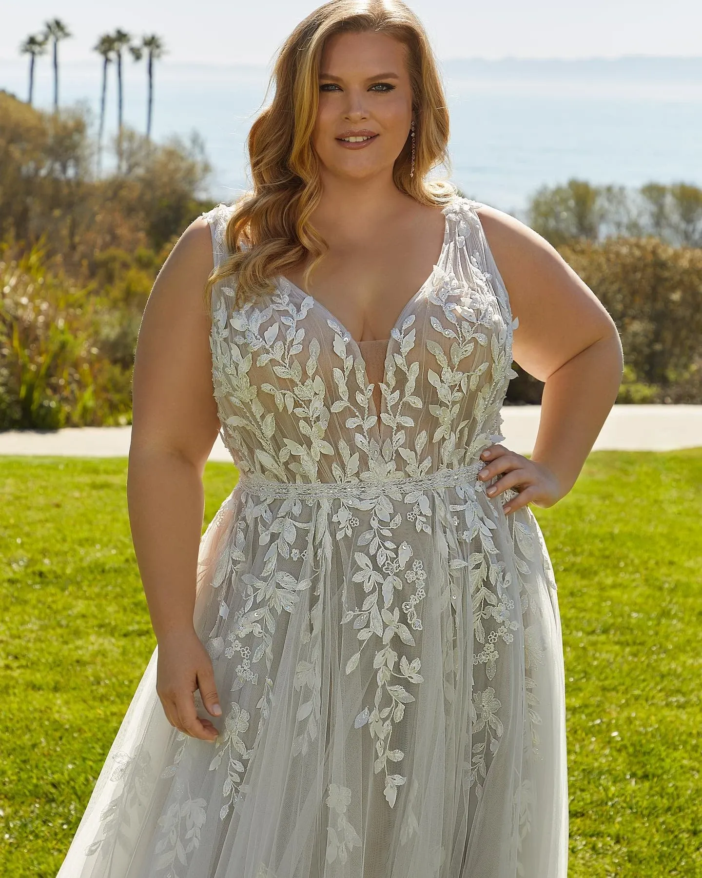 A bride wearing an elegantly designed wedding dress from Boutique MariClod, an esteemed vendor in the wedding dress category, stands outdoors. The gown features intricate floral lace patterns cascading across a sheer, flowing fabric, accentuating its romantic and stylish aura. The dress has a deep V-neckline and delicate straps, enhancing its graceful appearance. In the background, a serene view of tall palm trees and a distant ocean horizon adds a picturesque setting, perfectly complementing the bridal ensemble. This image showcases the exquisite craftsmanship and modern design offered by Boutique MariClod for brides in Canada.