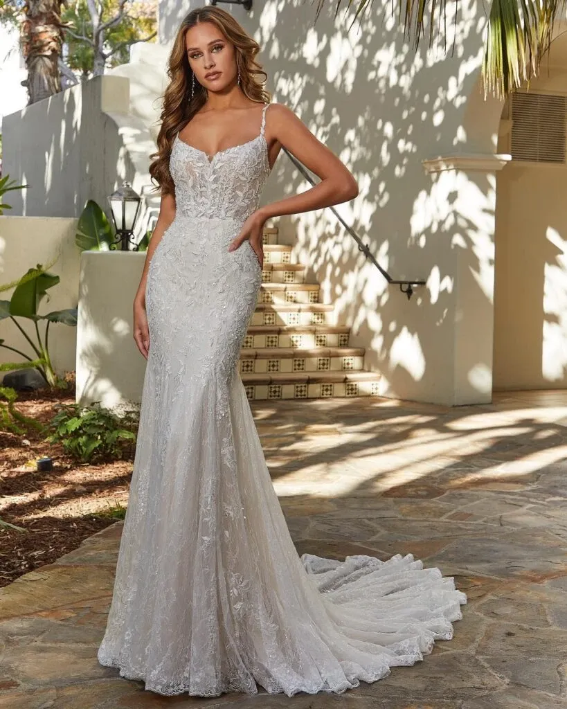 This image showcases a stunning wedding dress from Boutique MariClod, categorized under wedding dresses on Mariage Elegant, Canada. The dress features a form-fitting silhouette with a delicate lace overlay and elegant spaghetti straps. The gown's intricate lace detailing flows seamlessly into a graceful train. The bride stands outdoors on a sunlit stone pathway, framed by lush greenery and soft shadows, adding a natural, picturesque backdrop. This wedding dress exudes timeless elegance and sophistication, ideal for a modern bride seeking a classic look.