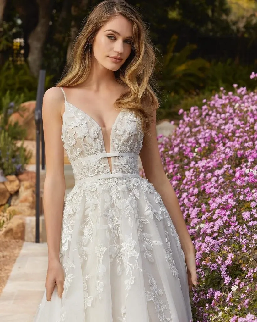 This image features a stunning wedding dress from Boutique MariClod, a vendor listed in the wedding dress category on Mariage Elegant, Canada. The dress showcases intricate floral lace patterns that embellish the bodice and flow elegantly onto the full skirt. The sleeveless design is complemented by delicate thin straps, and the slight V-neckline adds a touch of sophistication. The bride stands gracefully in a garden setting, surrounded by vibrant pink flowers and lush greenery, offering a romantic and picturesque backdrop perfect for a wedding celebration.