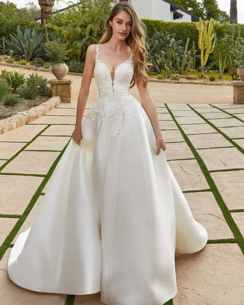 A bride is wearing an elegant wedding dress from Boutique MariClod, showcasing intricate lace detailing on the fitted bodice. The gown is sleeveless with delicate straps and features a plunging neckline that adds a touch of modern sophistication. The full skirt gracefully extends to the ground, creating a classic and timeless silhouette ideal for a wedding. The setting is an outdoor garden with stone pathways and lush greenery, providing a serene and romantic backdrop that complements the bridal attire. This image represents the Wedding Dress category for Mariage Elegant in Canada.