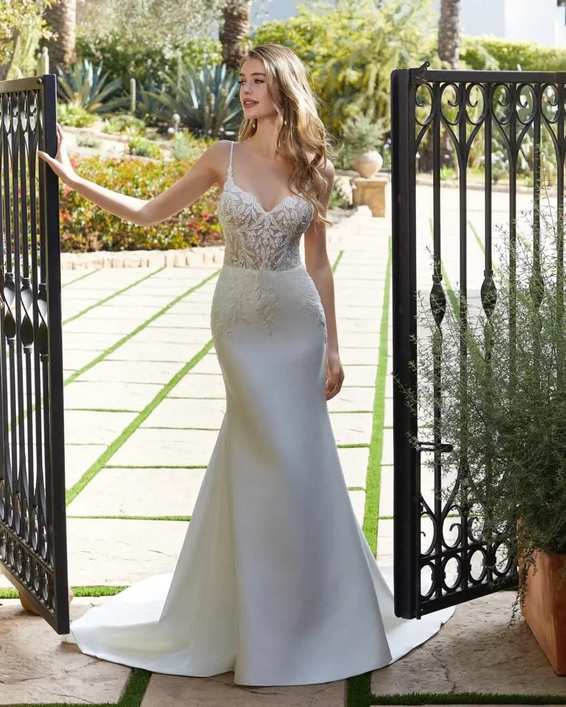 A bride stands gracefully in an elegant garden, showcasing a stunning wedding dress from Boutique MariClod, part of the Wedding Dress category at Mariage Elegant, Canada. The dress features intricate lace detailing on the bodice with delicate straps that complement the form-fitting silhouette, which gently flares into a classic train. Her loose, wavy hair adds a touch of romance to the overall look, perfect for a sophisticated wedding ceremony. The open wrought iron gate and lush greenery surrounding her create a picturesque setting, enhancing the dress's timeless elegance.
