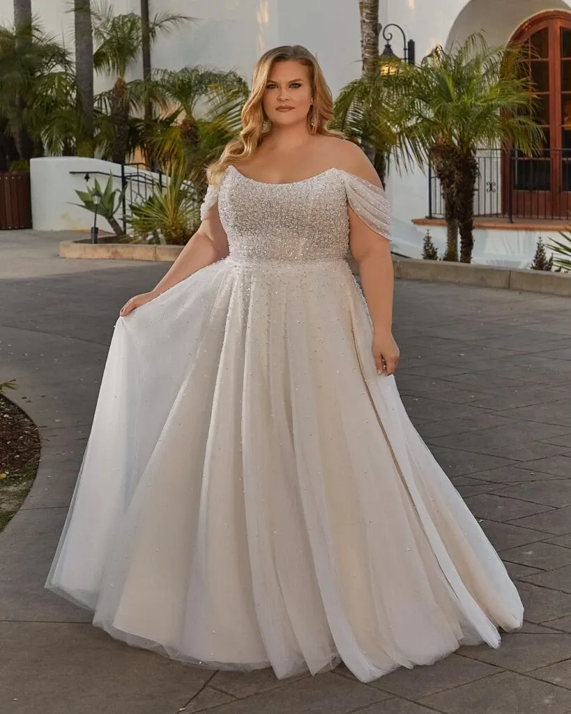 This image features a stunning wedding dress from Boutique MariClod, categorized under wedding dresses, showcased on Mariage Elegant, Canada. The dress elegantly flows in a full-length design with a delicate, sparkling overlay fabric. It has an off-the-shoulder neckline adorned with intricate beadwork, adding a touch of glamour. The setting includes a sunlit outdoor walkway lined with lush palm trees, creating a romantic atmosphere perfect for a wedding day. The model's long, wavy hair complements the dress beautifully, highlighting its sophisticated yet ethereal appeal.