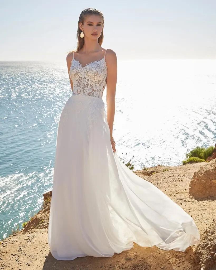 In this image from Boutique MariClod in the wedding dress category, a bride stands elegantly by a coastal backdrop. Her gown features a delicate lace bodice with intricate floral patterns and thin straps, flowing into a long, airy skirt that captures a sense of grace. The sunlight enhances the dress's beauty, creating a radiant effect, while the sparkling ocean serves as a stunning natural setting, ideal for a beach or seaside wedding. The scene encapsulates sophistication and serenity, reflecting the upscale offerings of Mariage Elegant in Canada.