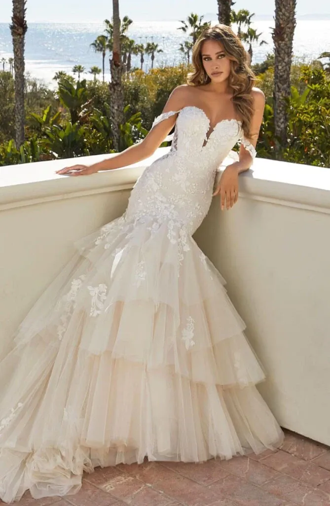 A bride models a stunning wedding dress from Boutique MariClod, featured in the Wedding Dress category. The gown showcases an elegant off-the-shoulder design with a sweetheart neckline, accentuating the intricate lace detailing. The fitted bodice flows into a voluminous, tiered tulle skirt, creating a classic and romantic silhouette. The setting appears to be a picturesque outdoor location, with palm trees and a scenic ocean view in the background, adding to the overall charm and sophistication of the image.