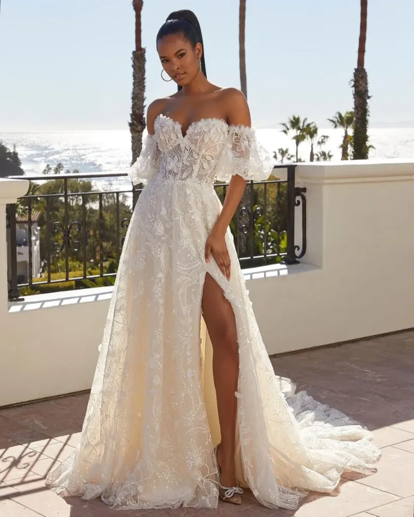 A bride modeling an elegant, off-the-shoulder lace wedding dress from Boutique MariClod, featured in the Wedding Dress category on Mariage Elegant, Canada. The gown showcases intricate lace detailing with a fitted bodice and a high slit that adds a modern touch. The backdrop features a sunny patio overlooking a scenic view of the ocean and palm trees, providing a romantic setting that complements the dress's elegance.