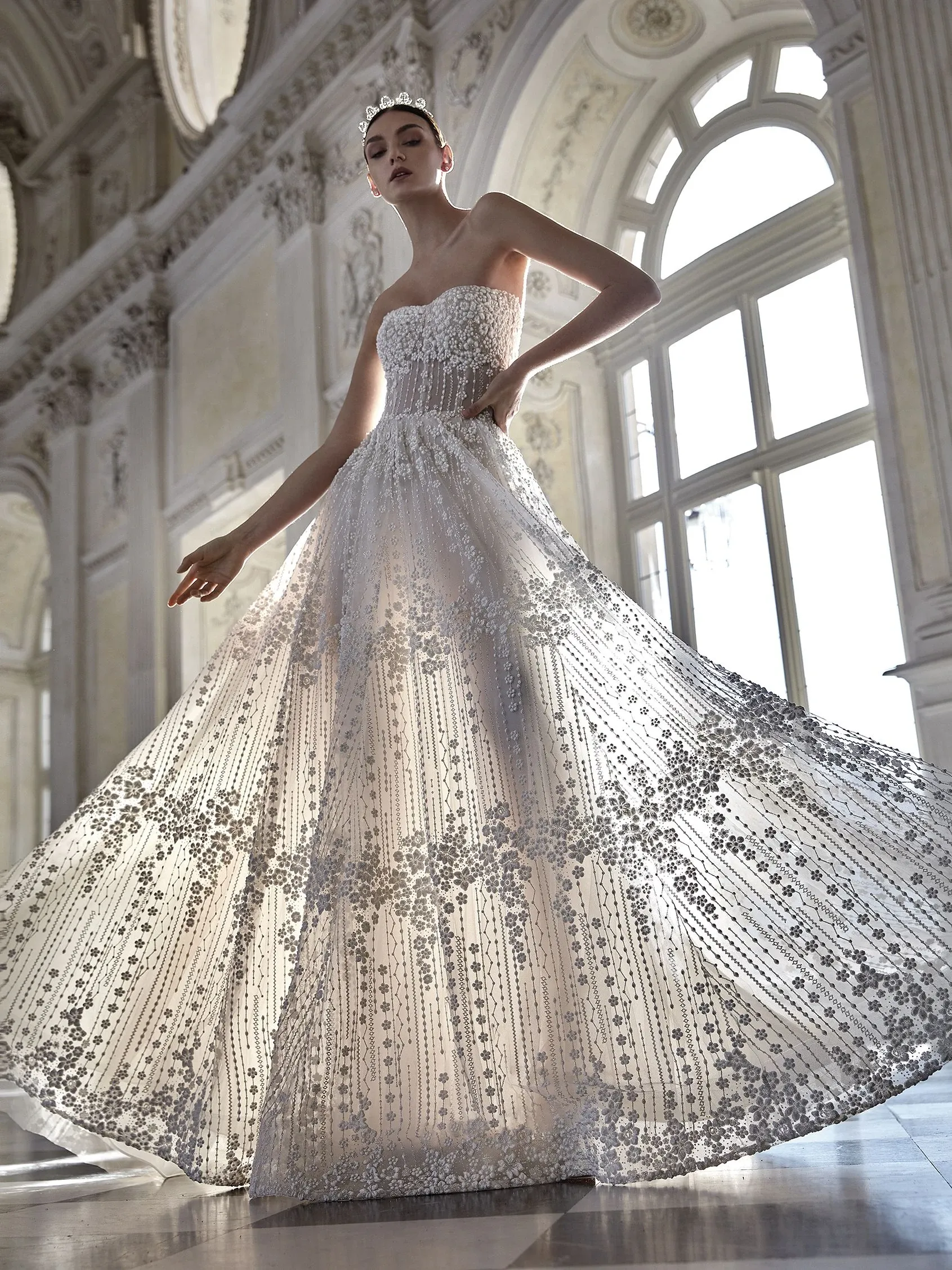 A bride elegantly poses in a grand, sunlit hall, showcasing a stunning wedding dress from Créations Vézina. The gown features intricate lace detailing with sparkling embellishments, creating an exquisite and timeless look. The strapless design and fitted bodice flow into a voluminous skirt, capturing the essence of sophistication and luxury. This masterpiece by Créations Vézina is perfect for a fairytale wedding, highlighting the beauty and craftsmanship expected in the wedding dress category. The majestic setting with tall windows and ornate architecture enhances the regal appearance of the ensemble, embodying the elegance of Mariage Elegant in Canada.