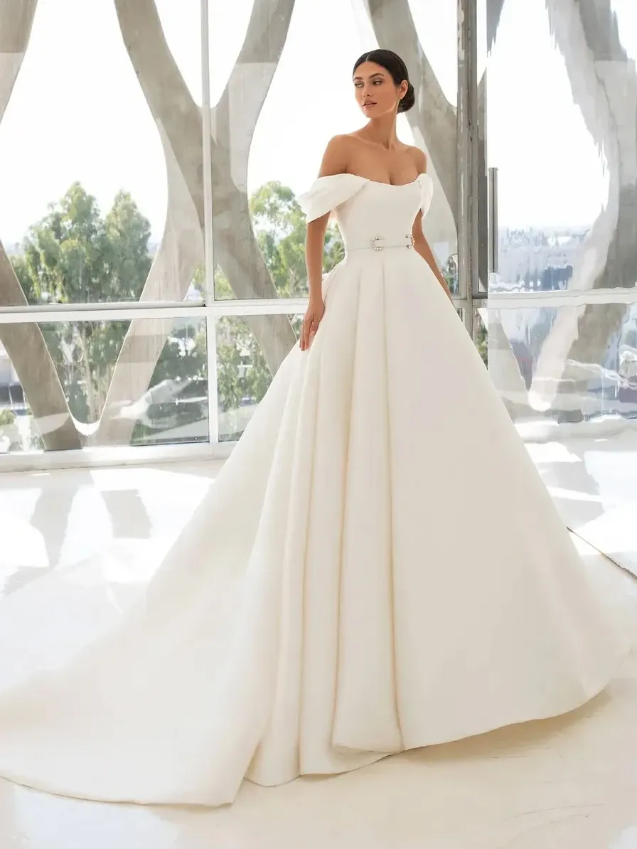 A bride is elegantly posing in a Créations Vézina wedding dress, showcasing the stunning design in this wedding dress category image. The dress features off-the-shoulder sleeves and a fitted bodice with intricate embellishments, flowing into a voluminous, pleated skirt. The background of large windows allows natural light to illuminate the sophisticated detailing of the gown, making it ideal for a classic and luxurious wedding setting in Canada.