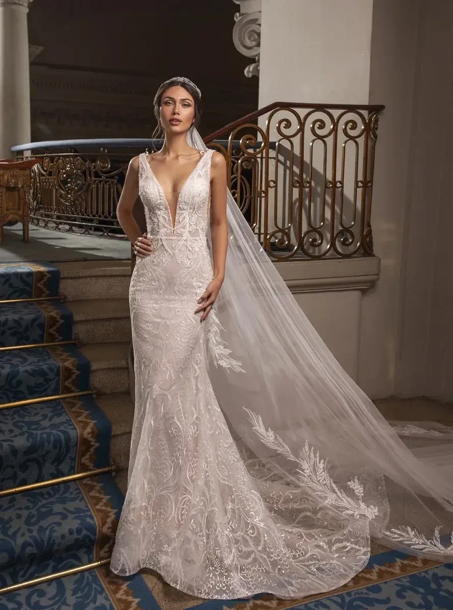 A bride stands gracefully on an ornate staircase, showcasing a stunning wedding dress by Créations Vézina in the wedding dress category. The dress features intricate lace detailing with a deep V-neckline and a form-fitting silhouette that elegantly flares into a subtle train. A long, delicate veil embellished with matching lace patterns cascades from her head, complementing the elegant surroundings. The setting, with its decorative iron railings and luxurious carpet, adds a sophisticated backdrop to this exquisite bridal attire.