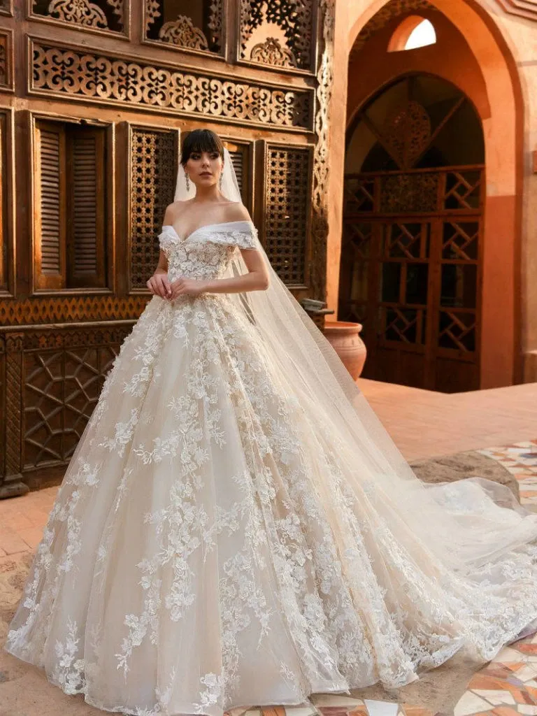 A bride is elegantly standing in a luxurious setting, showcasing a stunning wedding dress by Créations Vézina, featured in the Wedding Dress category. The gown is off-the-shoulder with intricate lace detailing and floral embroidery cascading down the voluminous skirt. The dress is complemented by a long, flowing veil. The background features ornate, carved wooden panels and an archway, enhancing the romantic ambiance suitable for a wedding. This image represents the exquisite craftsmanship and attention to detail that Créations Vézina brings to bridal fashion.