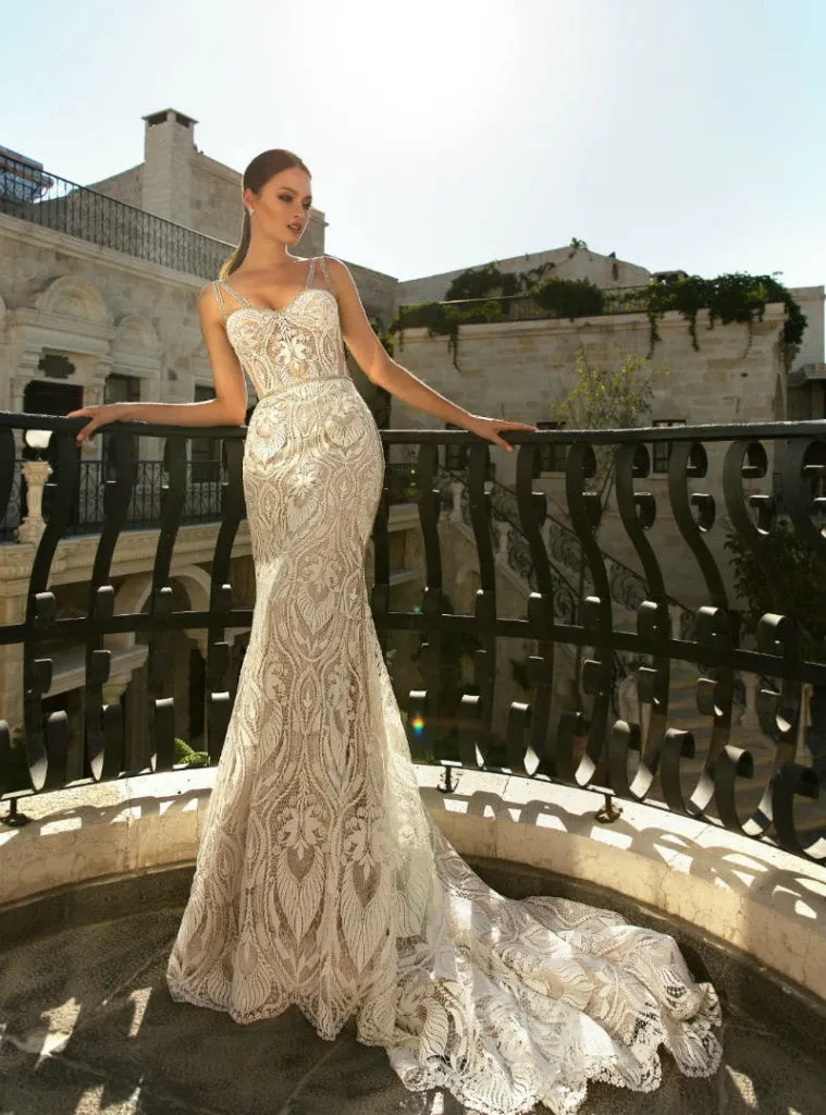 A bride elegantly poses on a stone balcony, wearing a sophisticated wedding dress by Créations Vézina, a vendor featured in the Mariage Elegant Canada wedding planning and vendor listing. The dress is an intricate lace design with a mermaid silhouette, fitted bodice, and flowing train, showcasing exquisite craftsmanship. The scene captures a classic and luxurious ambiance, perfect for an unforgettable wedding day.