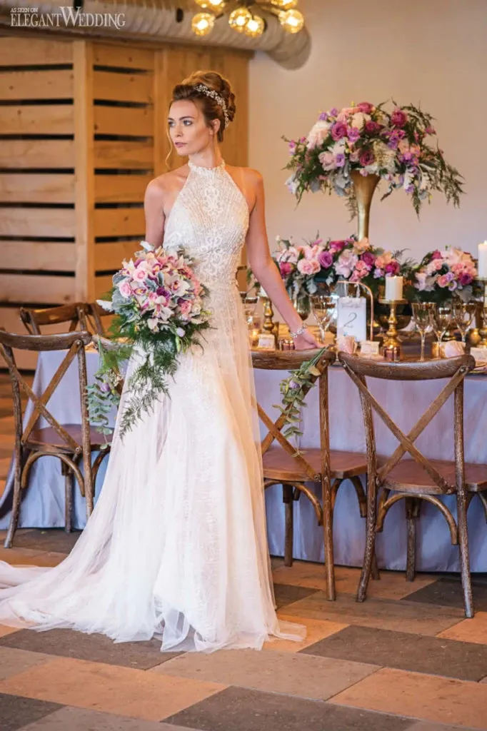 A bride stands elegantly beside a beautifully set wedding reception table, showcasing a stunning wedding dress by Créations Vézina. The dress features a high neck with intricate lace details and a flowing, sheer train. Her hair is styled in an elegant updo, complementing the sophisticated look. She holds a lush bouquet of pink and white flowers, mirroring the elaborate floral arrangements on the table. The table is adorned with candles, greenery, and floral centerpieces, creating a romantic and luxurious atmosphere, perfect for a wedding celebration. This image highlights the exquisite craftsmanship of the wedding dress, fitting seamlessly into a setting that exudes elegance and style.