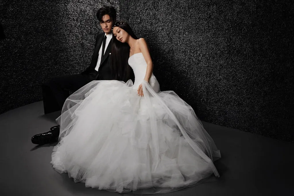 A bride and groom are seated against a textured dark wall. The bride is wearing a stunning strapless wedding dress from Créations Vézina in the wedding dress category. Her gown features a voluminous tulle skirt that elegantly drapes around her. The groom is dressed in a classic black suit, complementing the bride's attire. This image captures an intimate and stylish moment, showcasing the sophisticated and elegant bridal designs offered by Créations Vézina, ideal for weddings in Canada.