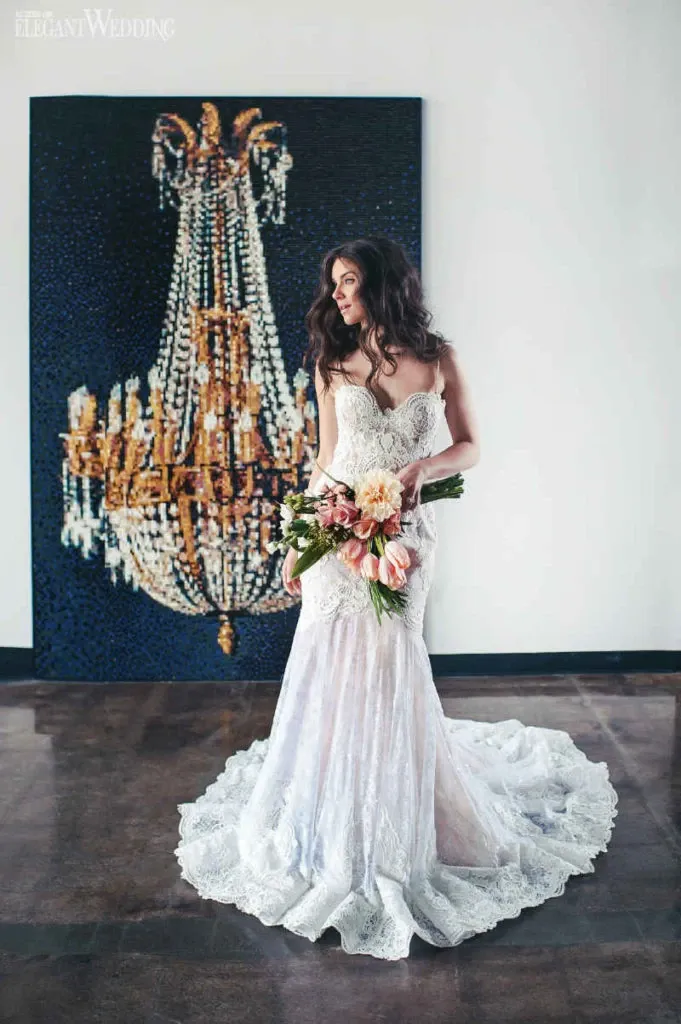 A bride wearing an elegant, strapless lace wedding dress by Créations Vézina stands gracefully in front of a large, artistic depiction of a crystal chandelier. The gown features intricate lace detailing and a flowing train that cascades onto the polished floor, enhancing its sophisticated design. She holds a bouquet of soft pink and peach flowers, adding a touch of color to the classic bridal look. This image represents the wedding dress category, ideal for couples seeking exquisite bridal fashion on the Mariage Elegant platform in Canada.