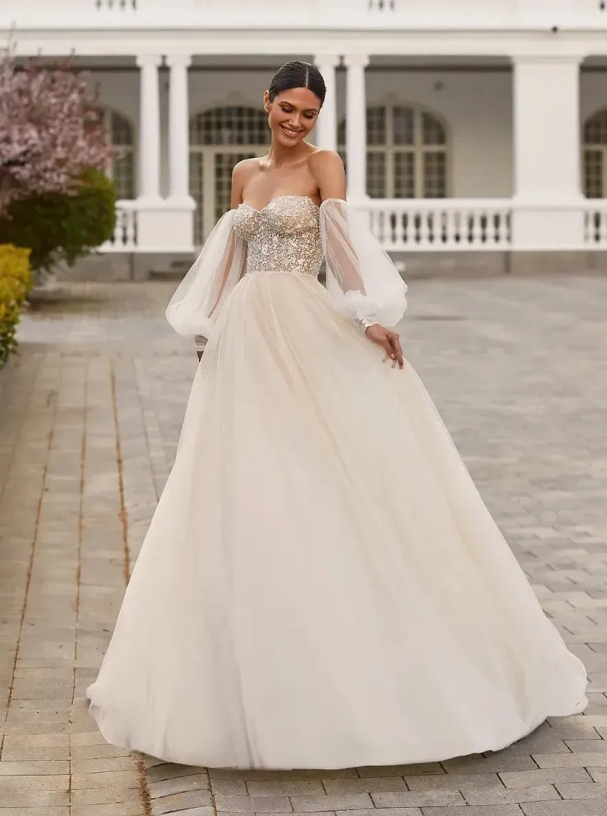 In this image for Mariage Elegant, a bride models an exquisite wedding dress by Créations Vézina, showcasing the vendor's elegance in bridal fashion. The dress features a fitted, sparkling bodice with intricate beading and off-the-shoulder tulle sleeves, adding a romantic and sophisticated touch. The flowing skirt cascades gracefully to the ground, made from layers of soft, ivory fabric. The setting is a picturesque outdoor venue with a cobblestone pathway and a beautifully maintained garden, providing a perfect backdrop for the stunning wedding attire in the Wedding Dress category.