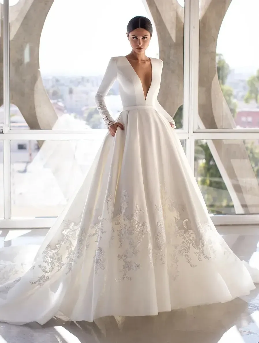 In this image, a model showcases a stunning wedding dress by Créations Vézina, falling under the wedding dress category for Mariage Elegant, Canada. The dress features a deep plunging V-neckline and long sleeves, exuding elegance. The full-length gown is adorned with intricate embroidery and subtle embellishments that catch the light. The bottom half of the dress flares out gently, with ornate lace details adding sophistication. The model stands in a spacious, light-filled room with large glass windows, highlighting the gown's elegance against the modern backdrop.