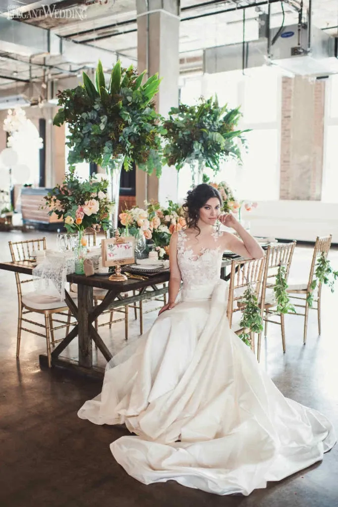 This image features a bride seated at a beautifully decorated table, showcasing a stunning wedding dress from Créations Vézina, a vendor listed under the Wedding Dress category on Mariage Elegant, Canada. The dress is elegant, featuring intricate lace detailing on the bodice and a flowing satin skirt. The setting is an airy, industrial-style venue with large windows and rustic elements. The table is adorned with lush floral arrangements and greenery, complemented by elegant tableware, contributing to a romantic and sophisticated wedding atmosphere.