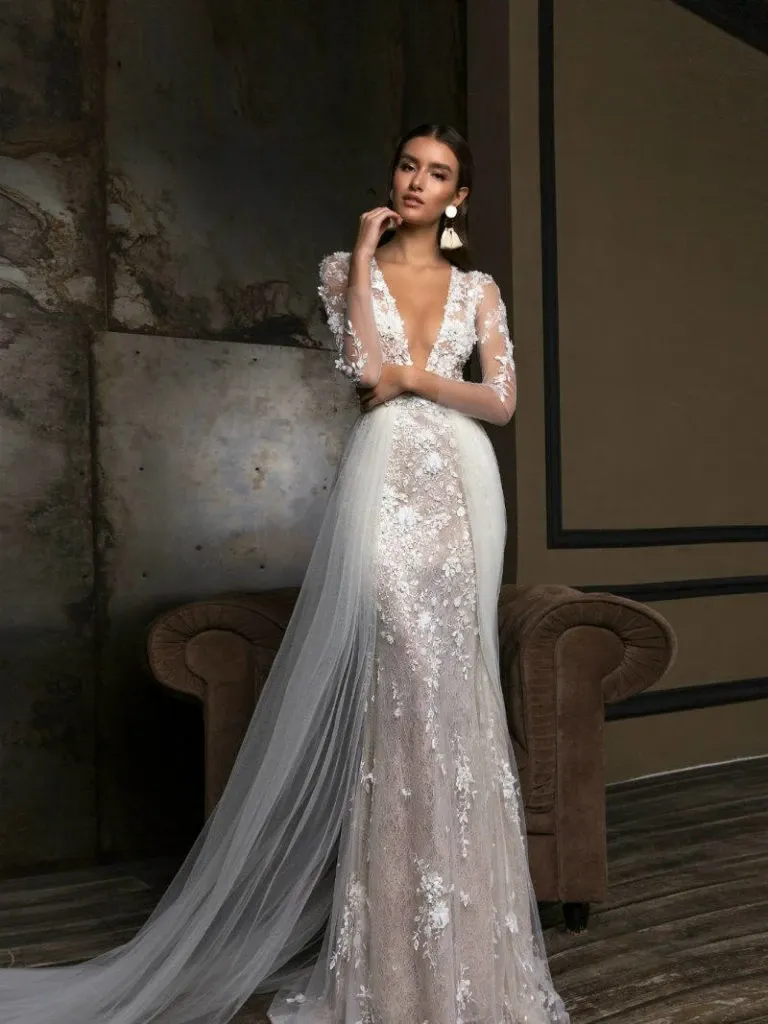 A stunning wedding dress by Créations Vézina, featured in the Mariage Elegant vendor listing under the wedding dress category, is showcased in the image. The gown is intricately designed with delicate lace appliqués and a deep V-neckline that exudes elegance. Long sheer sleeves adorned with floral lace patterns add a touch of romance, while the flowing tulle overlay enhances its ethereal beauty. The model poses gracefully against a sophisticated backdrop, highlighting the dress's exquisite craftsmanship and suitability for a modern, stylish wedding.