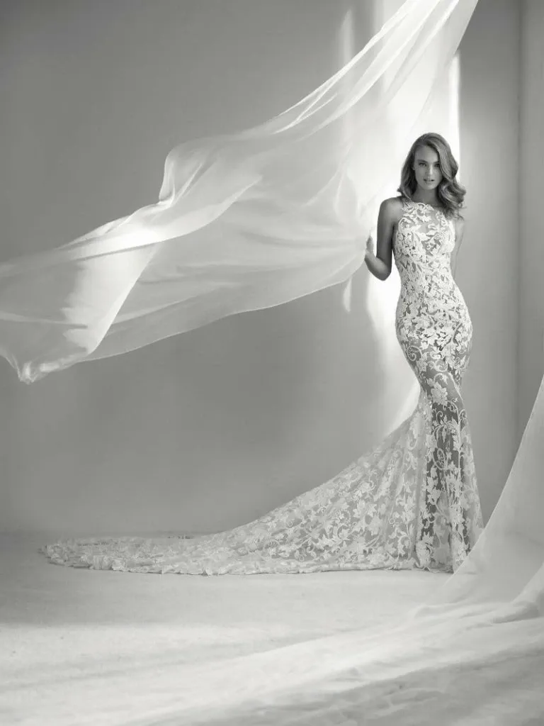 A bride stands gracefully in a fitted lace wedding dress by Créations Vézina, showcasing intricate detailing and elegance, perfect for a wedding setting. The gown features a form-fitting silhouette with a high neckline and an exquisite long train, highlighting the vendor's expertise in wedding dresses. Soft, flowing fabric drapes around her, adding a dreamy ambiance suitable for any elegant wedding.