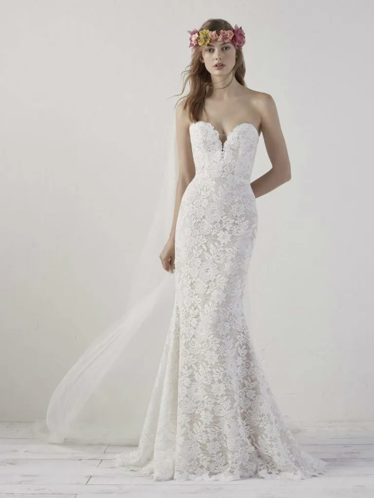 A bride models a strapless, form-fitting lace wedding dress designed by Créations Vézina, featured under the wedding dress category for Mariage Elegant in Canada. The gown, crafted with intricate lace detailing, showcases a sweetheart neckline and a slight mermaid silhouette that gently flares out at the knees. The model wears a floral crown with fresh blooms in soft pink and purple hues, complementing her long, wavy hair cascading over one shoulder. The background is minimalistic, highlighting the elegance and sophistication of the wedding dress.