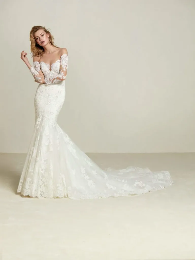 The image features a stunning wedding dress from Créations Vézina, showcased in the wedding dress category of Mariage Elegant, Canada. This exquisite gown displays a form-fitting silhouette with intricate lace detailing that extends through the bodice and down to the trailing hem. The dress has off-the-shoulder lace sleeves, adding a touch of elegance and sophistication. A soft, flowing train elegantly trails behind, completing the gown's romantic look. This design exemplifies the elegance and craftsmanship prized by brides seeking timeless wedding attire.