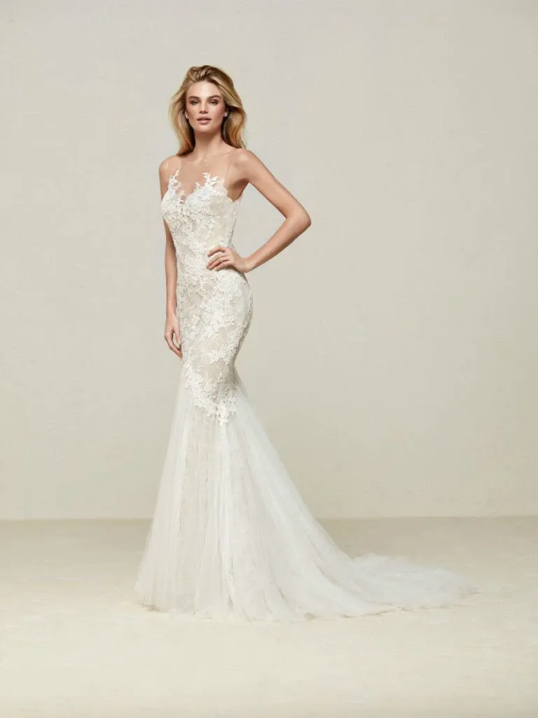 This image showcases a stunning wedding dress by Créations Vézina, a featured vendor in the Wedding Dress category on Mariage Elegant, Canada. The dress is a form-fitting, elegant gown with a mermaid silhouette, featuring intricate lace detailing and a sheer bodice. The delicate lace patterns extend partially over the shoulders and down the length of the train, creating a romantic and sophisticated look. The model poses gracefully, highlighting the gown's elegant design and craftsmanship, making it an ideal choice for a classic and refined wedding ceremony.