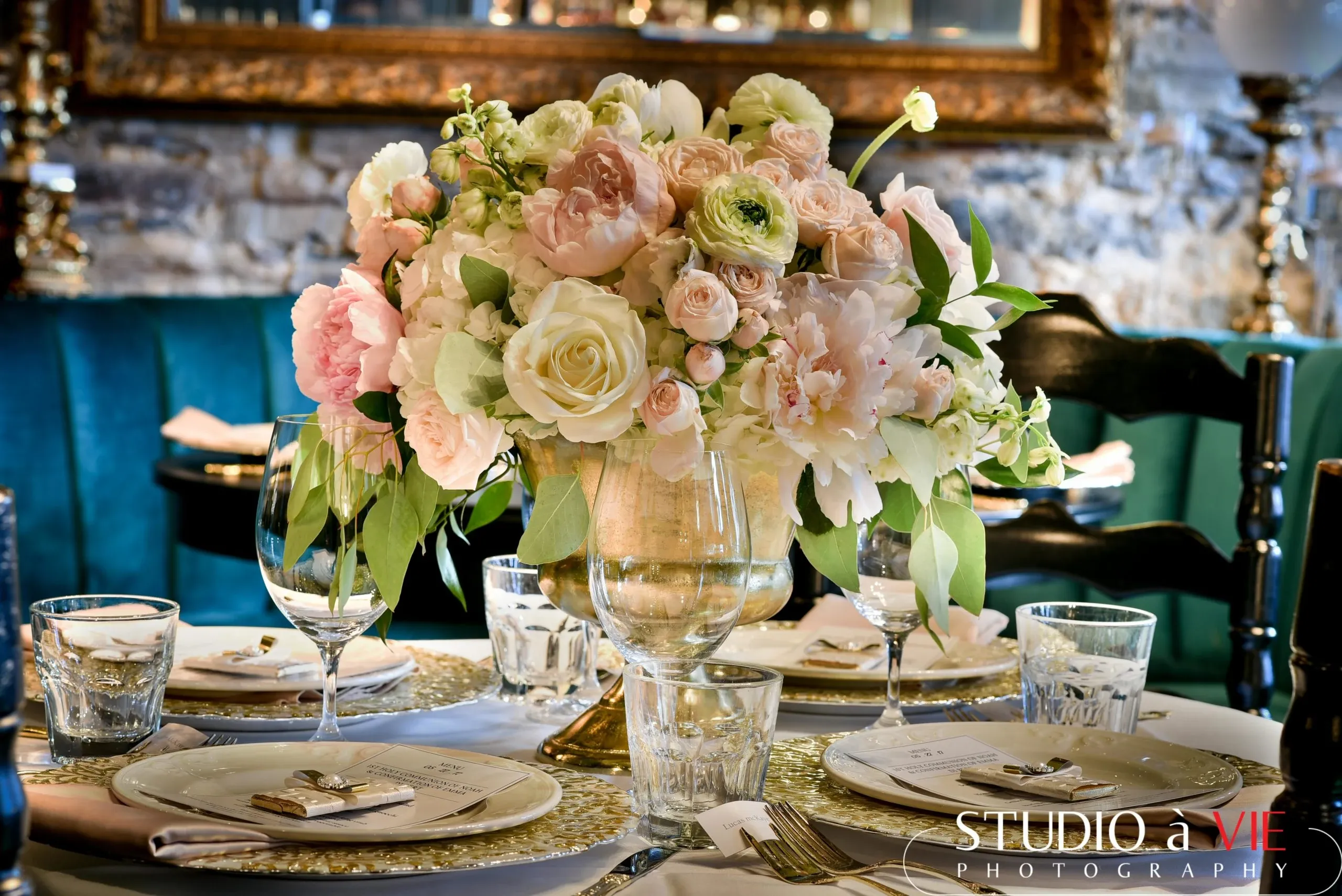 The image features a beautifully arranged wedding table centerpiece crafted by Lelili Fleurs, a vendor in the Wedding Florists category. This elegant floral arrangement showcases a stunning bouquet of pale pink and white roses, peonies, and ranunculus, accented by lush greenery. The flowers are placed in a clear glass vase, sitting atop a table set for a wedding reception. Surrounding the centerpiece are elegantly set plates with white napkins and glassware, all enhancing the sophisticated ambiance. The table is part of a warmly lit, upscale venue setting, suggesting a charming and romantic atmosphere suitable for a wedding celebration.