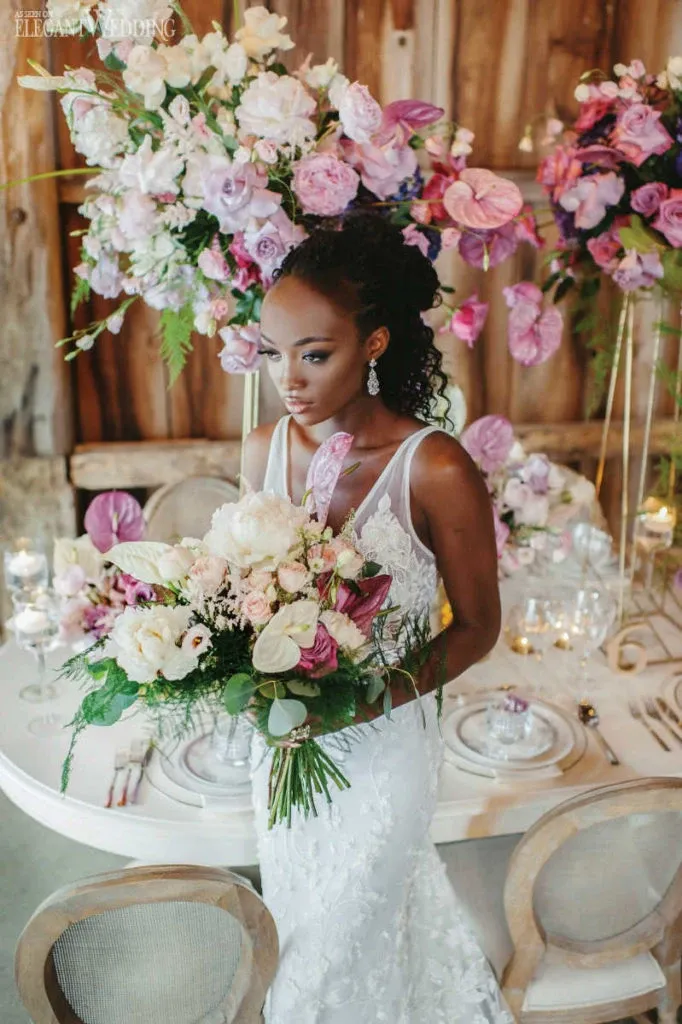 A bride stands elegantly in front of a beautifully arranged floral display by Lelili Fleurs, a featured wedding florist in Canada. The vibrant bouquet she holds is a blend of soft pinks, purples, and whites, complemented by lush greenery. In the background, two towering floral arrangements mirror the bouquet's color scheme, adding a romantic and sophisticated touch to the setting. The table is set with fine dinnerware, enhancing the overall elegance of the wedding day atmosphere. This image showcases Lelili Fleurs' expertise in creating stunning floral designs for weddings.