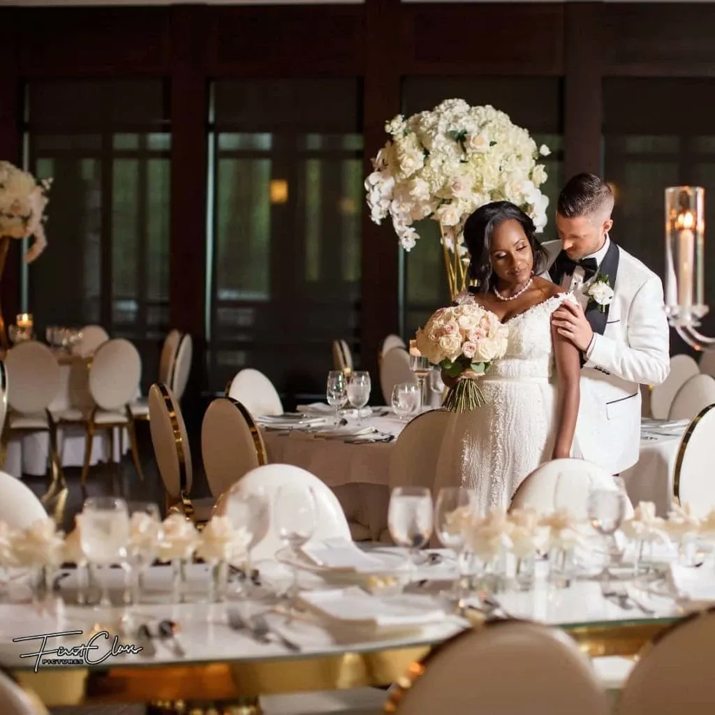 A elegantly decorated wedding reception room is shown, featuring a couple standing lovingly together. The bride holds a bouquet of white and blush roses by Lelili Fleurs, a wedding florist, while the groom, in a white tuxedo, embraces her gently. Behind them, beautifully arranged floral centerpieces consisting of tall, lush arrangements of white flowers are visible on the tables, adding a sophisticated touch to the setting. The tables are set with fine dinnerware and crystal glassware, reflecting the upscale ambiance suitable for a wedding celebration hosted by Mariage Elegant, Canada.