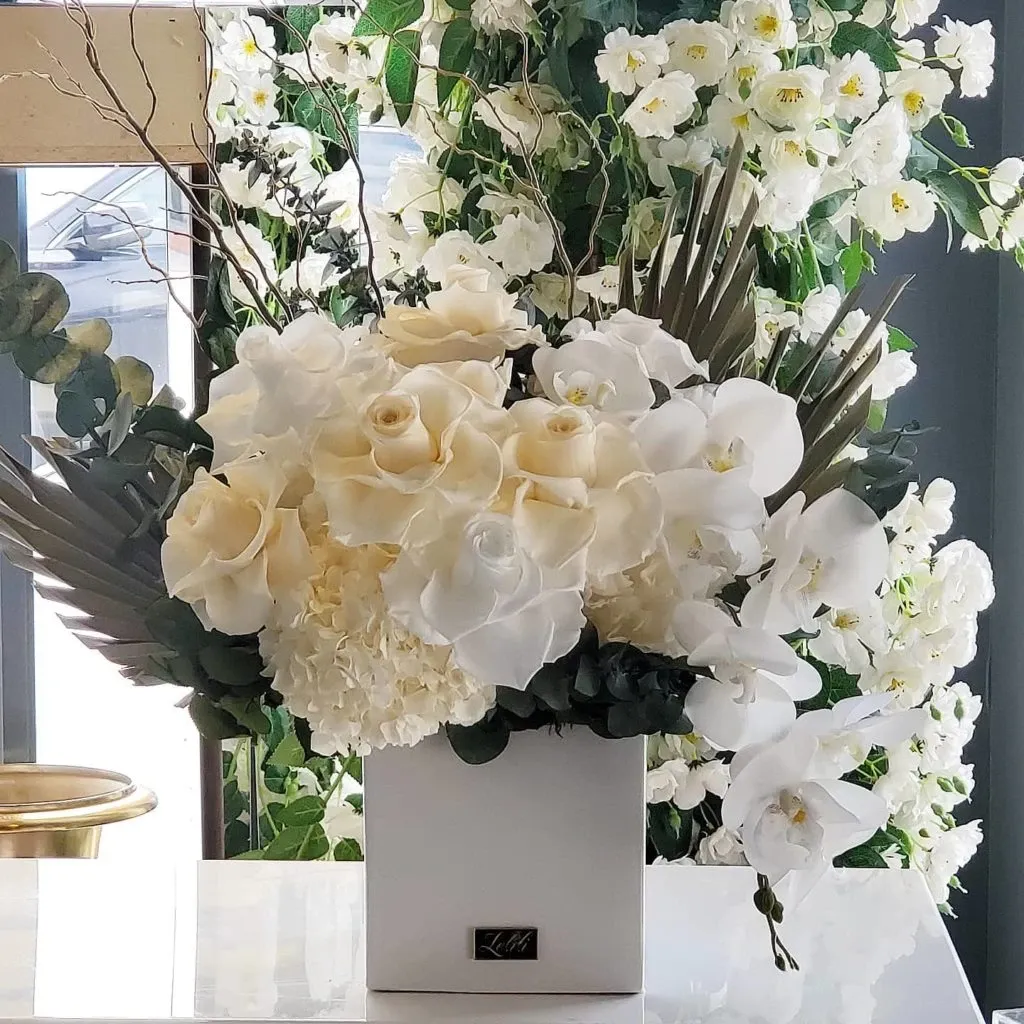 The image showcases an elegant floral arrangement by Lelili Fleurs, a wedding florist featured in the Mariage Elegant vendor list. The centerpiece consists of lush white roses, orchids, and hydrangeas, beautifully arranged in a modern white vase. Accents of greenery and slender branches add depth, creating a sophisticated and romantic display. The backdrop features additional cascading white flowers, enhancing the overall ethereal and luxurious theme ideal for weddings in Canada.