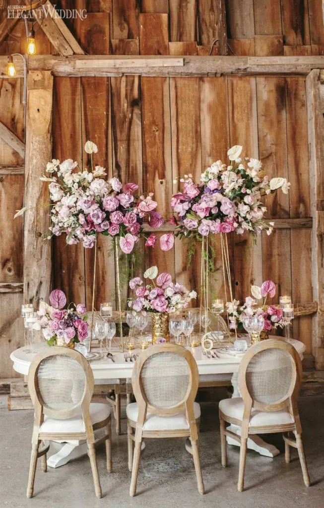 The image showcases a beautifully arranged wedding table setup by Lelili Fleurs, featured under the Wedding Florists category. The table is elegantly decorated with towering floral arrangements featuring a variety of pink and white flowers, including roses and anthuriums, set against a rustic wooden barn wall. The table is adorned with delicate glassware and candles, creating a romantic and sophisticated atmosphere ideal for a wedding setting. The overall decor emphasizes a harmonious blend of rustic charm and elegant floral artistry.