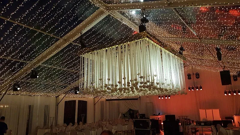 The image showcases an elegantly decorated wedding venue by Lelili Fleurs, categorized under Wedding Florists on Mariage Elegant, Canada. The focal point is a stunning floral installation hanging from the ceiling, featuring numerous white flowers cascading in a chandelier-like fashion. The venue is elegantly adorned with twinkling string lights, creating a romantic and enchanting atmosphere. The seating arrangement includes white draped chairs, complementing the sophisticated decor. This setup exemplifies the creative and exquisite floral artistry provided by Lelili Fleurs for weddings.