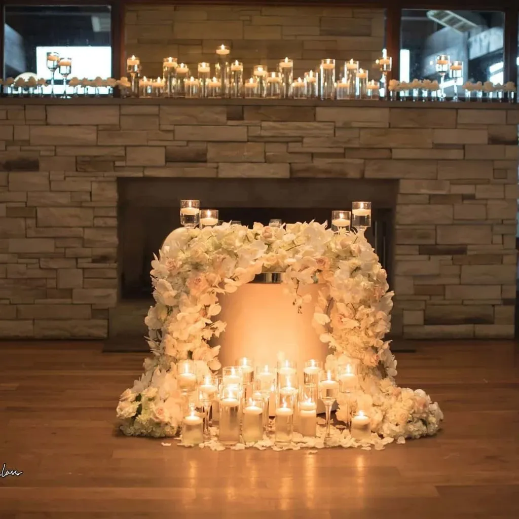 The image showcases an elegant wedding floral arrangement by Lelili Fleurs, categorized under Wedding Florists. It features a stunning arch of white and pale pink flowers, including roses and lilies, beautifully arranged around the base. In the center, numerous candles of varying heights are placed, creating a warm, romantic ambiance perfect for wedding ceremonies or receptions. Behind the floral display, a rustic stone backdrop enhances the sophisticated and inviting atmosphere. The scene is set indoors, with additional rows of candles visible on a ledge above, adding to the overall elegant and serene setting ideal for a memorable wedding event.