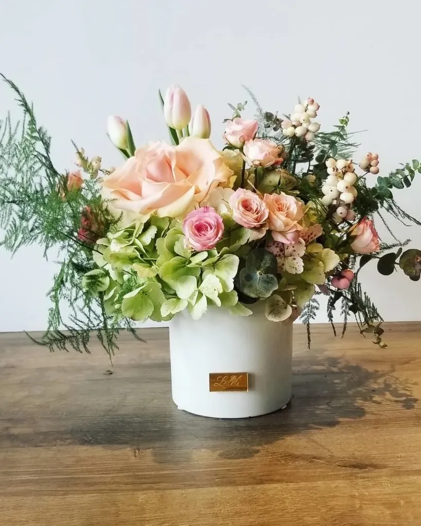 A beautiful floral arrangement by Lelili Fleurs, a wedding florist in the Mariage Elegant vendor listing in Canada. The arrangement features a mix of delicate pink tulips, large blush roses, and small pink spray roses. These are complemented by pale green hydrangeas, white berries, and lush greenery, all artistically arranged in a sleek white vase with a gold Lelili Fleurs label. This elegant floral design is perfect for enhancing the ambiance of a sophisticated wedding setting.