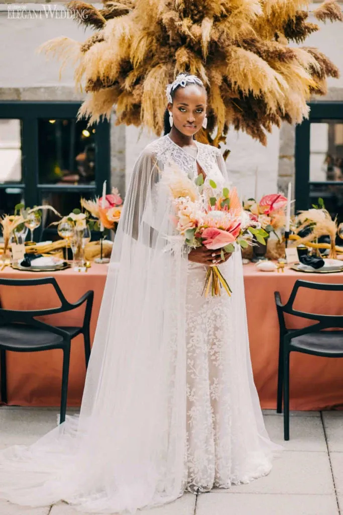 A bride stands elegantly in front of a beautifully set wedding reception table adorned with vibrant floral arrangements. She wears a detailed, lace wedding gown with a flowing cape, holding a bouquet featuring pink and coral flowers. Behind her, a stunning large arrangement of pampas grass adds a dramatic backdrop. Lelili Fleurs, a wedding florist, has crafted these exquisite floral designs, ideal for an elegant and stylish wedding setting. This image captures the sophisticated and creative flair of Lelili Fleurs in contributing to a memorable wedding ambiance.