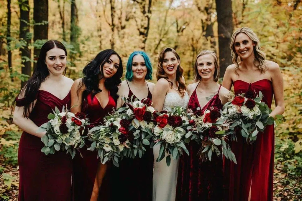 In this image for the wedding florists category, provided by Lelili Fleurs, six bridesmaids are standing in a wooded area surrounded by autumn foliage, creating a picturesque backdrop. They are all holding beautifully arranged bouquets featuring deep red, white, and blush flowers with lush greenery, showcasing the elegant floral design typical of wedding settings. The bridesmaids are wearing coordinated dresses in shades of burgundy and wine, enhancing the sophisticated, romantic theme suitable for a fall wedding. This image reflects the artistry and attention to detail expected from Lelili Fleurs, a featured vendor in the Mariage Elegant wedding planning service in Canada.