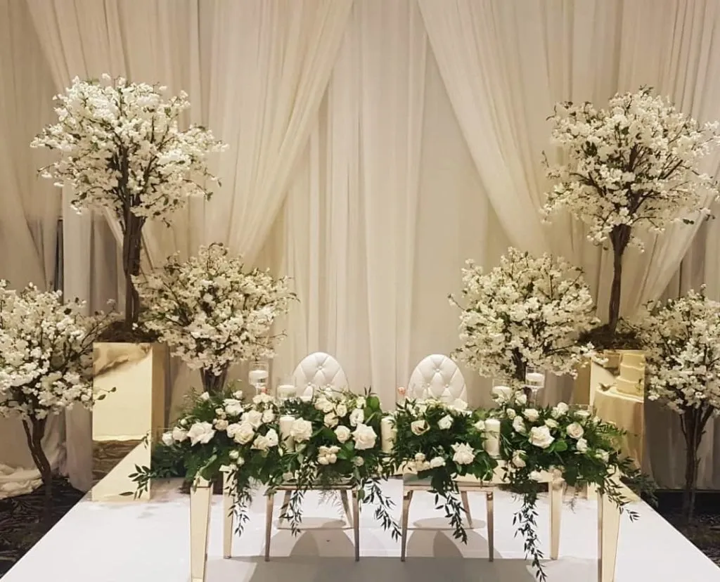The image showcases a stunning wedding floral arrangement by Lelili Fleurs, categorized under wedding florists at Mariage Elegant, Canada. The setting features an elegantly decorated bridal table adorned with lush white roses and greenery, perfectly accentuating the sophisticated atmosphere. Behind the table, tall, meticulously arranged floral trees with delicate white blossoms stand against a backdrop of sheer white drapes, creating a romantic and graceful ambiance ideal for a wedding celebration. The two plush white chairs add a touch of elegance, completing this breathtaking floral setup.