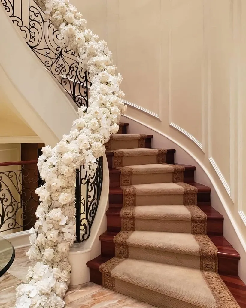 The image showcases an elegant spiral staircase adorned with a luxurious garland of white flowers, crafted by Lelili Fleurs, a wedding florist featured on Mariage Elegant, Canada. The floral arrangement gracefully follows the curve of the ornate wrought-iron railing, adding a touch of sophistication and romance to the setting. The staircase is covered with a traditional, richly patterned carpet, complementing the classic wedding decor theme. This setup exemplifies the artistry and attention to detail provided by Lelili Fleurs in creating stunning floral displays for weddings.