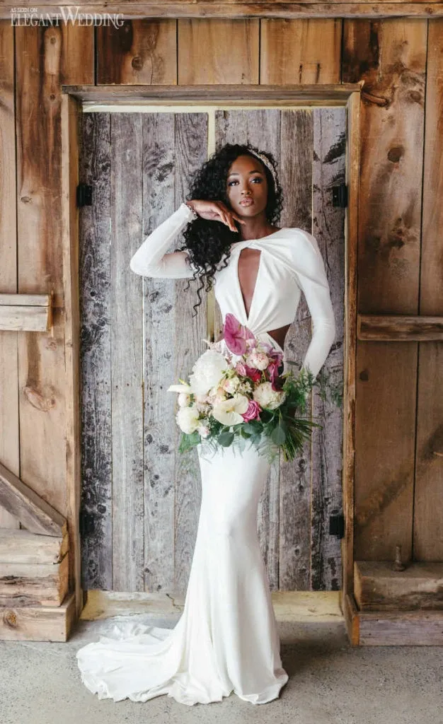 A bride stands elegantly in front of a rustic wooden door, showcasing a stunning bouquet from Lelili Fleurs, a Wedding Florist vendor at Mariage Elegant, Canada. She wears a sleek, white gown with a dramatic cutout neckline, complementing her poised pose. The bouquet features a harmonious blend of pink, white, and green floral arrangements, adding a touch of romance and sophistication to the scene. The image captures the essence of rustic elegance, ideal for wedding inspiration.