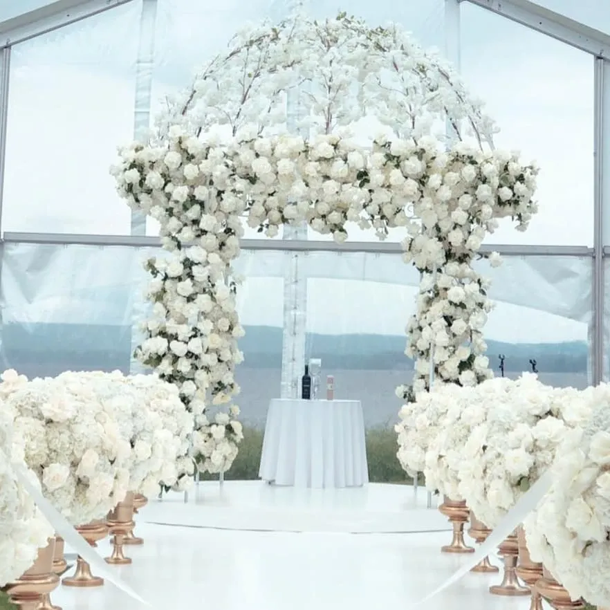 The image showcases a stunning floral arrangement by Lelili Fleurs, a wedding florist featured on Mariage Elegant in Canada. Displayed is a beautifully decorated wedding arch covered entirely in lush white flowers, including roses and hydrangeas, set against a transparent marquee tent. The aisle is elegantly lined with matching arrangements on gold stands, creating a luxurious and romantic atmosphere. In the background, a table is prepared for the ceremony, enhancing the sophisticated setting perfect for a wedding celebration.