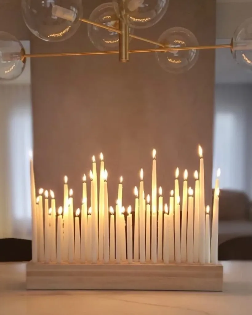 This image showcases an elegant display by Lelili Fleurs, a renowned wedding florist. The arrangement features a variety of white taper candles, artfully placed at different heights in a wooden holder, creating a romantic and serene ambiance. The soft glow of the candlelight reflects beautifully, casting warm illumination ideal for a wedding setting. Overhead, a modern, spherical light fixture adds a touch of sophistication, complementing the overall aesthetic. This display is a perfect example of how a skilled wedding florist like Lelili Fleurs can enhance the atmosphere of a wedding venue with creative floral and decor elements.